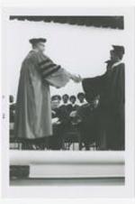 A man and a woman, wearing graduation caps and gowns, shake hands on stage at commencement.