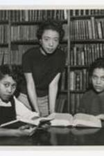A group portrait of three women at a table in front of a large bookshelf.