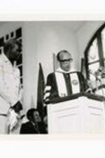 President Hugh Gloster speaking at podium with Zambian President Kaunda and unidentified person. Written on verso: Zambian President Kaunda receives Honorary Degree---Commencement 1978 Ebenezer Baptist Church.