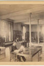 Interior of the Graves Library in Stone Hall with male students looking at books while a woman sits at a desk. Written on verso: B. F. Allen in front