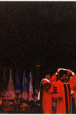 Men in regalia stand on a stage during a program and hood C. Eric Lincoln.