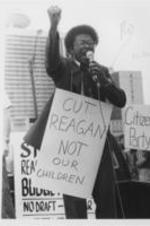 Reverend Timothy McDonald is shown speaking with a microphone during a protest against the Reagan administration in Atlanta, Georgia. Written on verso: Coalition Against Reagan -- Rev. Timothy McDonald, assistant pastor of Atlanta's Ebenezer Baptist Church, rallies a crowd of demonstrators outside an Atlanta hotel during a visit by President Reagan. The group was protesting Reagan's economic policies and military buildup at the expense of the poor.