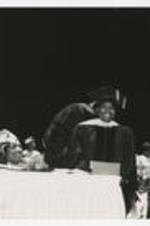 A view of a young woman, wearing graduation cap and gown, holding a diploma on stage at commencement.