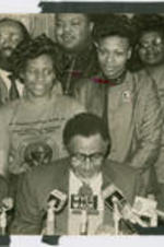 Southern Christian Leadership Conference (SCLC) President Joseph E. Lowery is shown speaking at a press conference related to SCLC's boycotts of Winn-Dixie stores with his wife Evelyn sitting to his right (at left in photo) and SCLC staff members behind him. Two individuals (Brenda Davenport and Kim Miller) wear a shirt marking the first national holiday for the birthday of Martin Luther King, Jr.