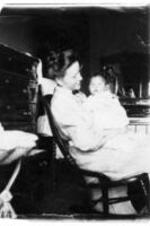 A woman sits in a chair holding a baby. Written on verso: Mrs. Towns and Helen.