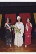 A women, wearing a long-sleeve floor-length white and gold dress with matching head wrap, sash and flower bouquet, poses with two other women, wearing formal dresses with tiaras and sashes, on stage.