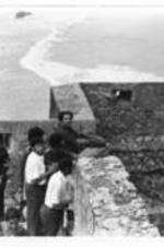 A group of men standing by a battlement and looking off the edge where men bring in a boat.