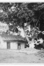 A small building with a woman standing in the doorway. Written on verso: Kitchen and site of old dormitory. Garraway, Liberia.