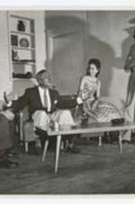 View of three men and a woman on stage seated on sofa, view of living room furniture.