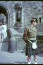 An unidentified man in a suit talks to a man in a kilt in front of a castle entrance.