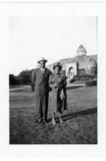 An unidentified couple stand on the grass in front of a large glass building.