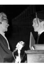 Grace Towns Hamilton stands at a podium shaking hands with a man wearing a cap and gown. Written on verso: John Spencer? A.U. Ch. [illegible] 9. T.H-Oct. 16-1981.