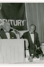 An unidentified man speaks at a podium alongside two other men and a woman seated.