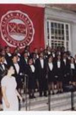 A group of men and women, wearing suits with bow ties, sing on steps of a building under a banner "Clark Atlanta University."