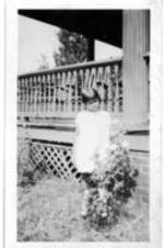 A young girl stands outside of a house and examines a rose bush.