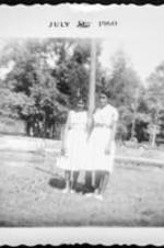 Two unidentified women wearing white dresses lean on a pole.