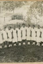 A group of young women graduates wearing white dresses and holding diplomas stand in the corner of a yard. A Universal Pictures movie poster of "The Doomed Battalion" is in the distance on a building exterior.