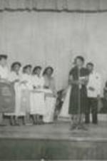 An unidentified group stands on a stage. One woman holds a sign that reads "Food Value Charts".