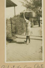 A girl carries a basket on her shoulder and walks through a garden gate. Written on recto: Girl at the open gate.