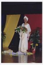 A women, wearing a long-sleeve floor-length white and gold dress with matching head wrap, tiara and sash, stands on stage holding a flower bouquet.