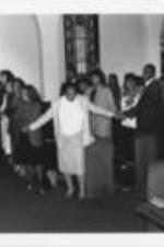 Evelyn G. Lowery (fourth from left, first pew) is shown holding hands and singing with others during SCLC/WOMEN's World AIDS Day event at Cascade United Methodist Church in Atlanta, Georgia. Written on verso: SCLC/WOMEN's World AIDS Day 1990