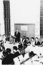 Alumni sit at a large banquet table as one man stands and talks.