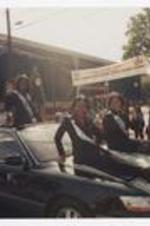 Three women, wearing a tiaras and sashes, sit on the hood and stand in the sun roof of a black car on road in front of campus gate.