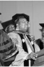 C. Eric Lincoln, wearing regalia, stands at a podium and receives a hood in a ceremony.