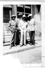 A group of unidentified men in suits stand on the sidewalk in front of a shoe shine shop and a lawyer's office.