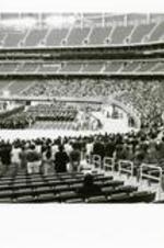 View of stadium, stage, and crowd at commencement. Written on verso: Commencement, 1996; The class of 1996 reached an all-time high of 466 graduates. An overflow crowd of 9,000 parents and friends showed up at Cathedral of the Holy Spirit/Chapel Hill Harvestor Church for their commencement (7,000 seat capacity). Myrlie Evers-Williams, chair of the National Boards of Directors of NAACP delivered the address.
