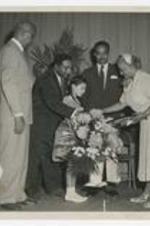 A young girl holding a bouquet of flowers on stage with three men and a woman.