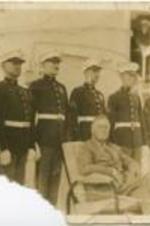 President Franklin Roosevelt sits in a chair on deck while six naval officers stand behind him.