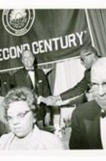 Hugh Morris Gloster shaking hands with another man at apodium. Banner in background reads "The Second Century."
