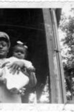 An unidentified man in uniform stands on a porch, holding a small baby.