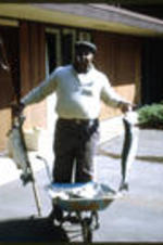 C. Eric Lincoln stands outside of a house holding two large fish and a fishing pole.