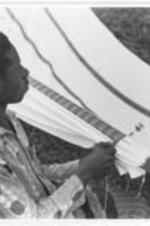 A young man stitches white cloth onto a hammock.