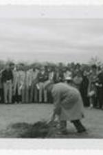 Man shovels dirt at a groundbreaking ceremony with spectators in background.