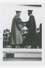 Two men, wearing graduation caps and gowns, shake hands on stage at commencement.