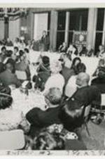 Men and women sit at dining tables with a man speaking at podium.