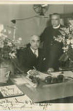 William Thompkins sits at his desk holding a pen while an unidentified man stands behind him. Written on recto: To my friends; Mr. and Mrs. Irvin H. McDuffie. From William J. Thompkins.
