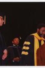 Colin Powell watches as Thomas W. Cole, Jr. and another man stand at the podium at the summer commencement.