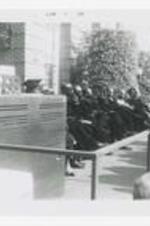 Men and women, wearing graduation caps and gowns, sit next to piano on an outdoor stage at commencement.
