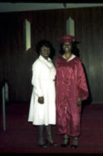 An unidentified young woman wearing a red cap and gown stands next to an unidentified older woman, possibly her Grandmother.