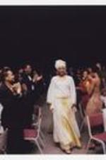 A women, wearing a long-sleeve floor-length white and gold dress with matching head wrap, walks through an audience of people standing at round dining tables.