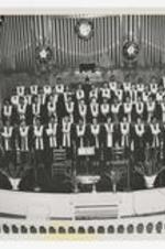 Indoor group portrait of choir on stage, view of pipe organ in background.