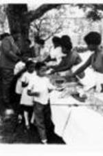 Unidentified women serve food to attendees at a cookout.