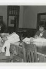 Two young women sit at a desk with fabric, a spool of thread, and other sewing items in a classroom.