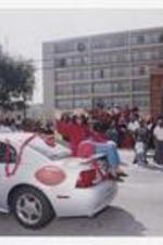 A woman, wearing a red shirt and hat, sits on the back of a silver car decorated with balloons, stars, football decal and sign "CAU Staff Assembly Homecoming '02" on street with parade audience in background.