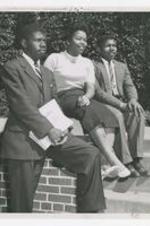A young woman and two men sit on a low brick wall next to stairs.