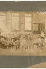 Children and their teachers sit in a circle in a classroom. Written on verso: Miss Gertrude H. Ware, kindergarten children